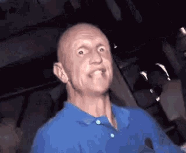 a bald man in a blue shirt is making a funny face while sitting in a dark room .