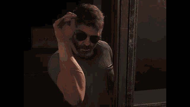 a man wearing sunglasses and a beard is standing in front of a door and waving his hand .