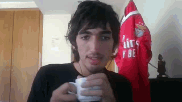 a man is holding a cup of coffee in front of a red shirt that says " airs y be "