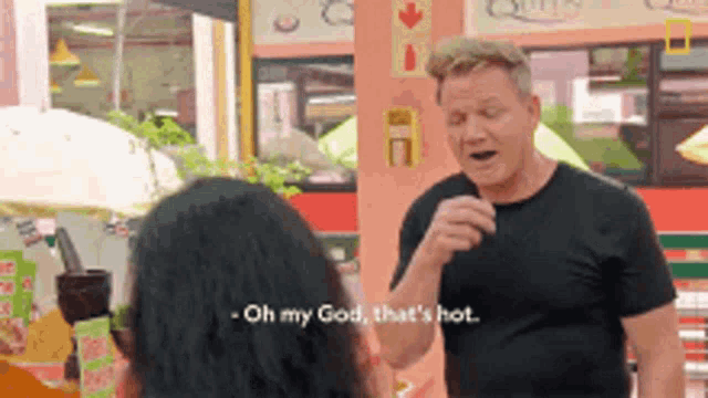 a man in a black shirt is talking to a woman and says oh my god that 's hot .