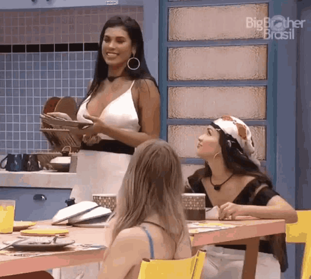 three women are sitting at a table in a kitchen with big brother brasil written on the wall behind them .