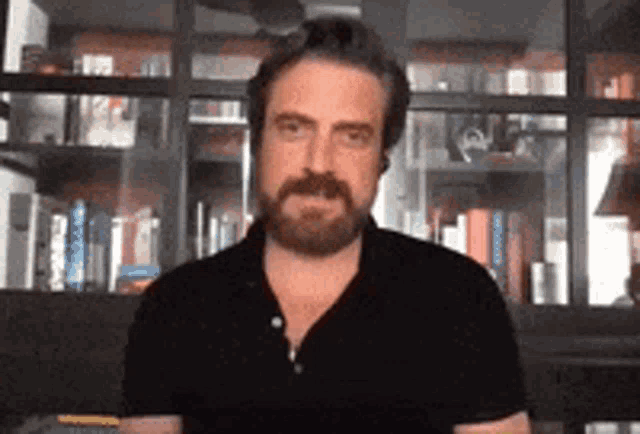 a man with a beard is standing in front of a bookshelf and looking at the camera .