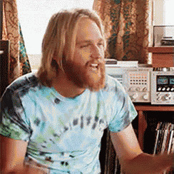 a man with long hair and a beard wearing a tie dye shirt is laughing .