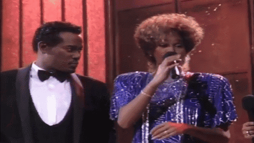 a woman in a blue dress is singing into a microphone while a man in a tuxedo looks on .