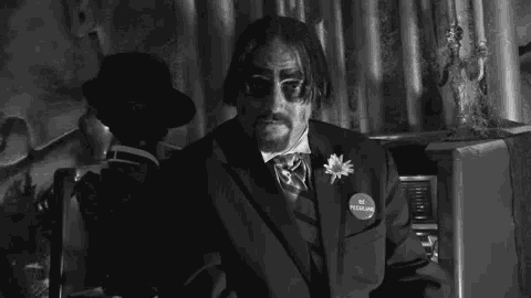a black and white photo of a man in a suit and tie sitting in a room .