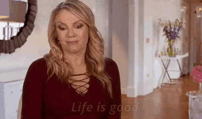 a woman in a red shirt is standing in a living room with the words `` life is good '' written on the wall .