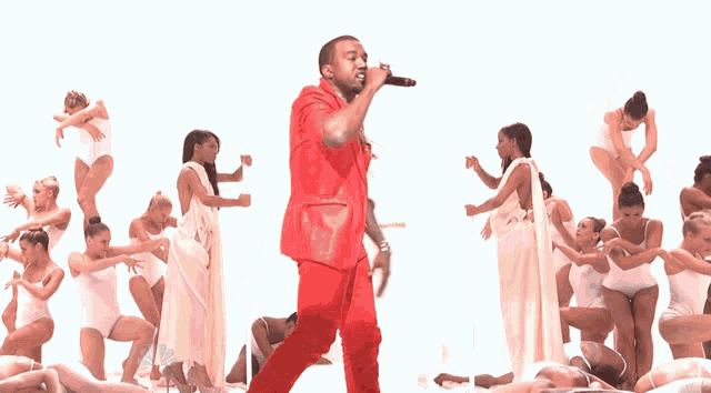 a man in a red suit is singing into a microphone in front of a group of women in white dresses