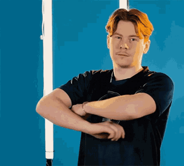 a young man in a black t-shirt is standing with his arms crossed and looking at the camera .