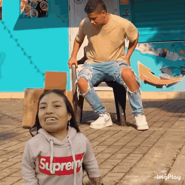 a woman wearing a supreme hoodie is standing next to a man in a chair