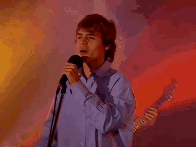 a young man in a blue shirt is singing into a microphone while holding a guitar .