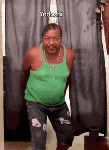 a man wearing a green tank top and ripped jeans is standing in front of a curtain with yodoshi written on it .
