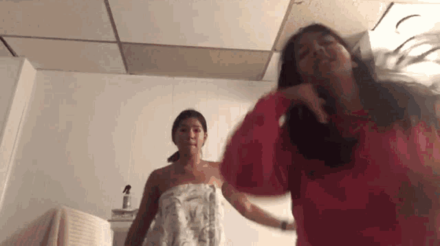 two women are standing next to each other in a bathroom . one of the women is wrapped in a towel .
