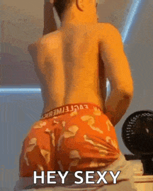 a shirtless man in orange underwear is sitting on a bed next to a fan and says hey sexy .