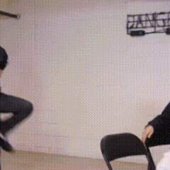 a man is standing in front of a white wall while another man sits in a chair in a room .