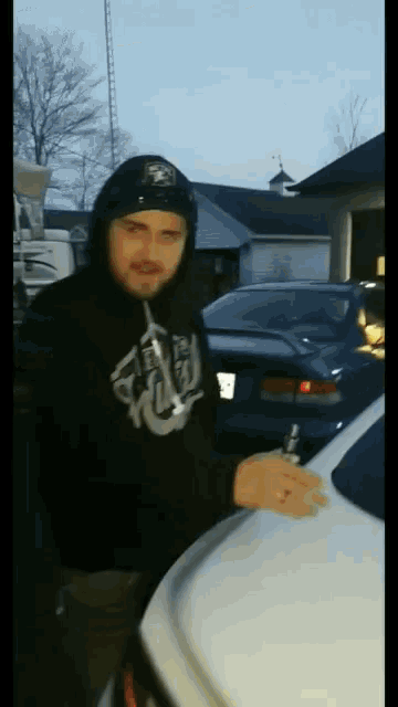 a man wearing a black hoodie with a skull on it is standing next to a car