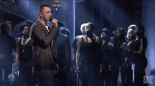 a man singing into a microphone with a snl logo on the bottom right