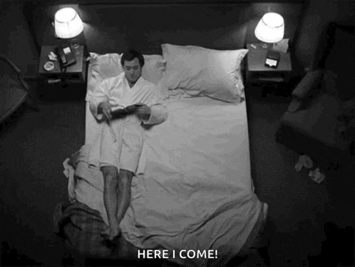 a man in a robe is laying on a bed reading a book and says here i come .