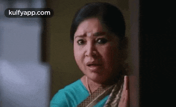 a woman in a blue shirt is standing in front of a door and making a funny face .