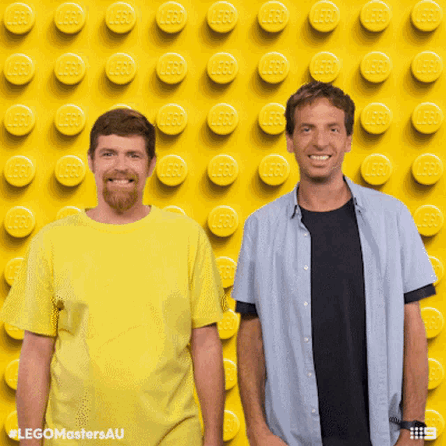 two men standing in front of a wall of yellow lego blocks