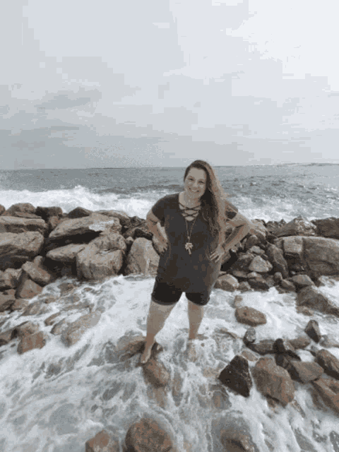 a woman stands on a rocky beach with her feet in the water