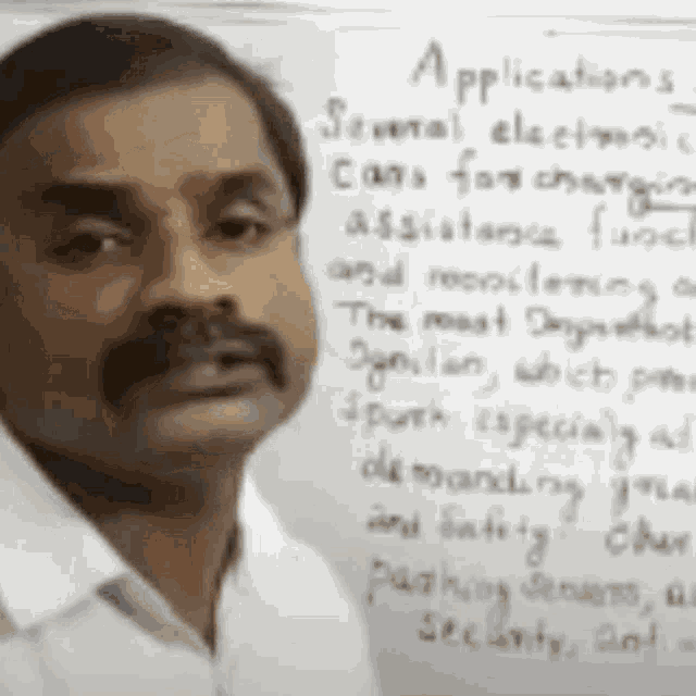 a man with a mustache is standing in front of a white board with handwriting on it .