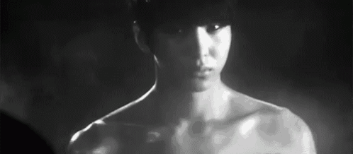 a man without a shirt is standing in the dark and looking at the camera in a black and white photo .