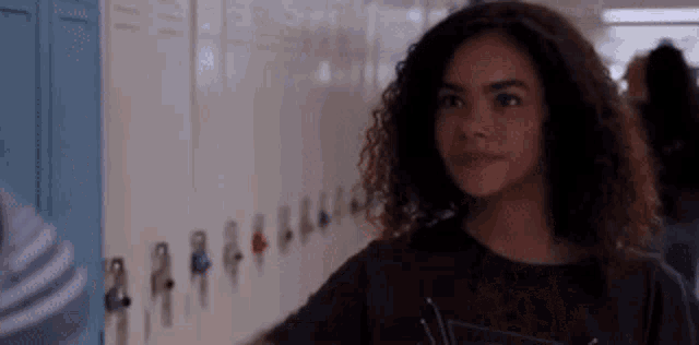 a girl with curly hair is standing in front of a row of lockers in a hallway .