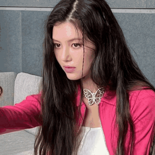 a woman with long black hair wearing a pink jacket with a butterfly necklace