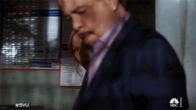 a man in a suit and a woman in a purple shirt are standing in front of a nbc poster