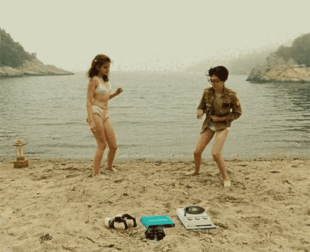 a woman in a bikini is dancing on the beach next to a man in a plaid shirt
