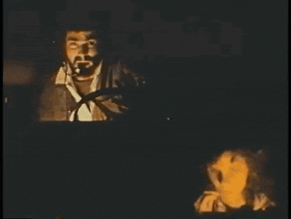 a man with a beard is smoking a cigarette in a dark room while a woman looks on .