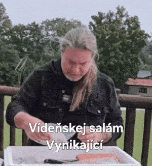 a man with long hair and a beard is sitting at a table with a container of food in front of him .