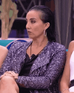 a woman in a sequined jacket and earrings is sitting in a chair with her hands folded .