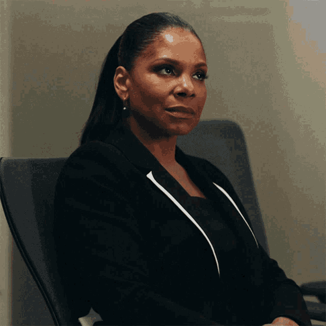 a woman in a black suit sits in a chair and looks to the side