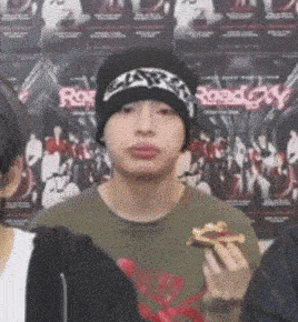 a man wearing a beanie is holding a slice of pizza in front of a poster that says roadway