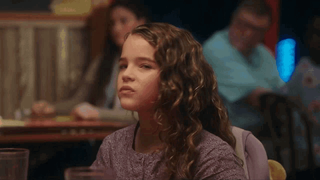 a little girl with curly hair is sitting at a table with a glass of water