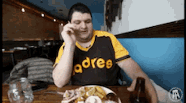 a man in a padres jersey sits at a table with a plate of food