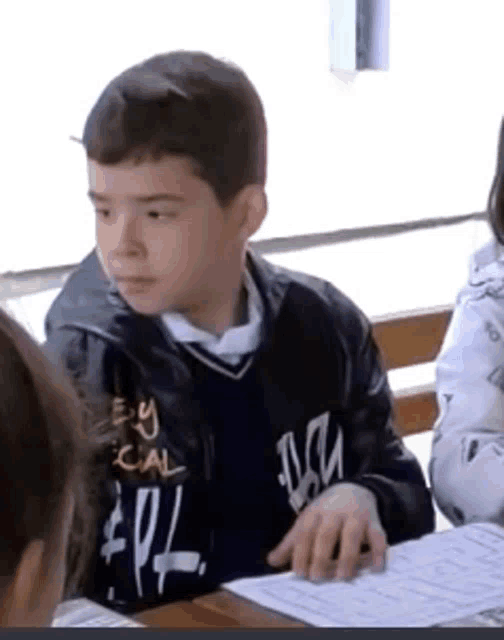 a boy wearing a black jacket that says " by social " sits at a desk