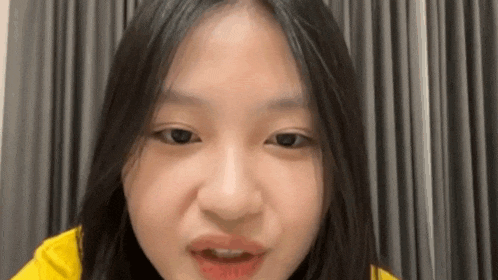 a young girl with long black hair is making a funny face in front of a gray curtain .