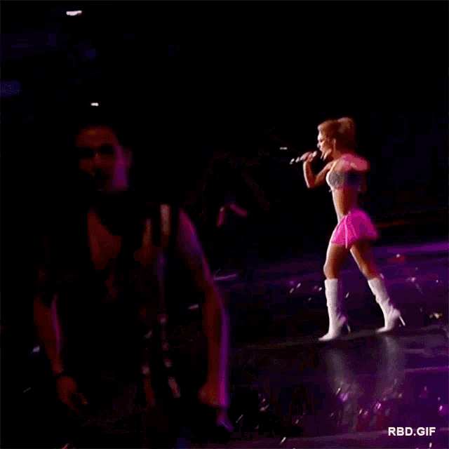 a man is holding a microphone on a stage with a woman in a pink dress behind him ..
