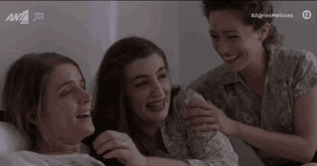 three women are laughing in a bed with the ant logo behind them