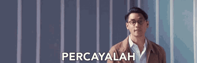 a man wearing glasses and a brown jacket is standing in front of a wall with the words percayalah written on it