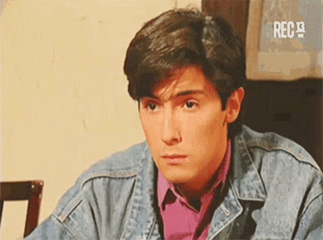 a young man wearing a denim jacket and a pink shirt is looking at the camera .