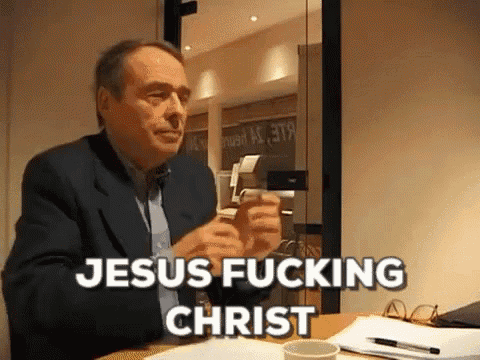 a man in a suit is sitting at a table and says jesus fucking christ .
