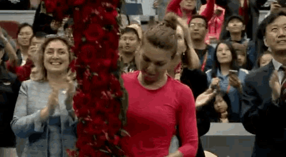 a woman in a red shirt is standing in front of a crowd of people holding a bouquet of flowers .
