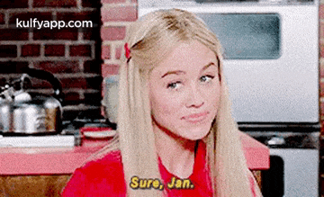 a blonde woman in a red shirt is sitting in a kitchen talking to a man .