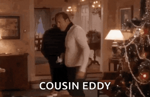 a man is standing in a living room next to a christmas tree and a lamp .