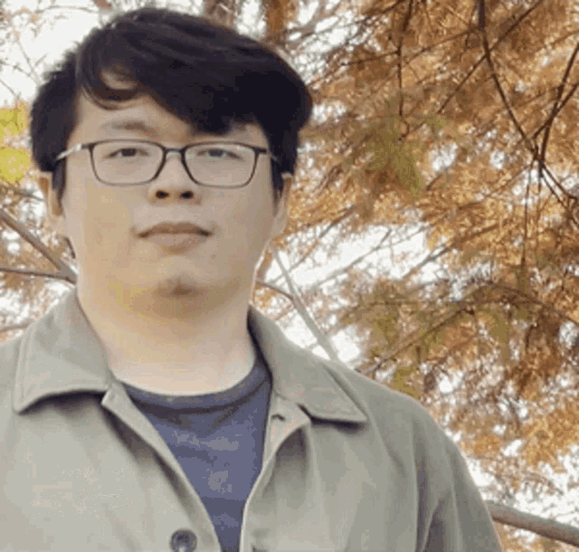 a man wearing glasses and a green jacket stands in front of trees