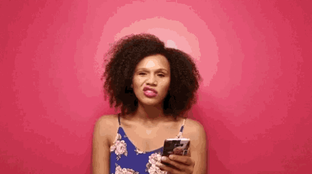 a woman with curly hair is holding a cell phone in front of a pink background .