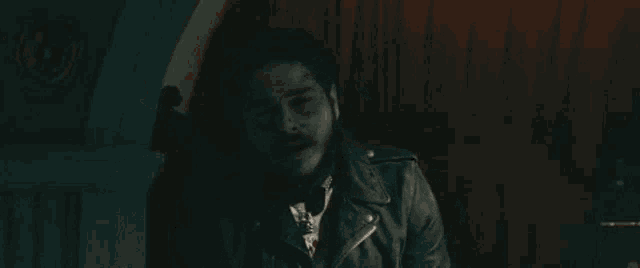 a man with a beard is wearing a leather jacket and smoking a cigarette in a dark room .